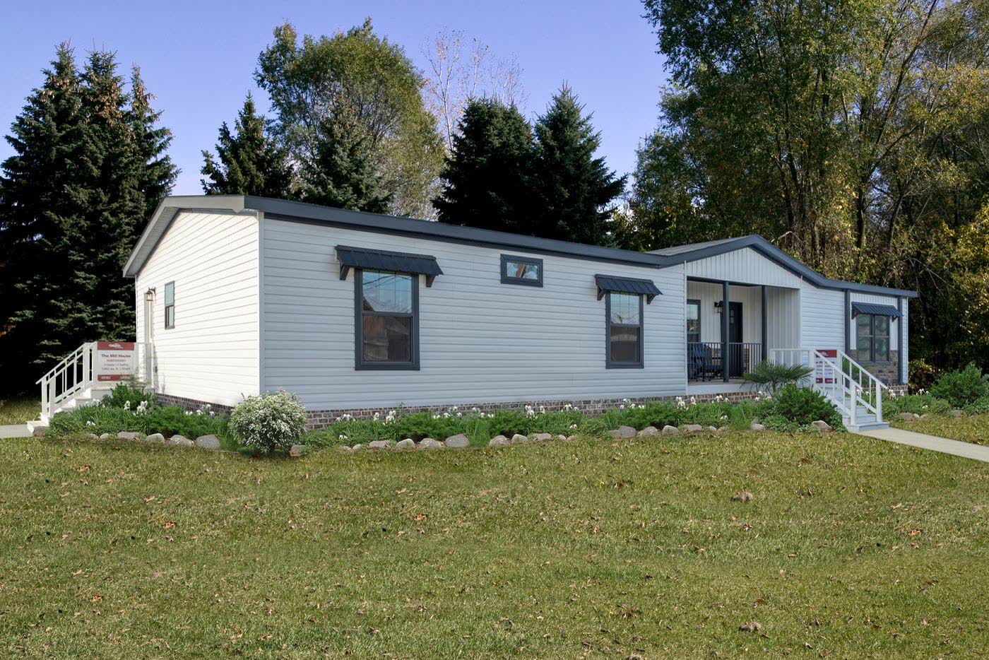 A white manufactured home with a covered porch among tall trees.