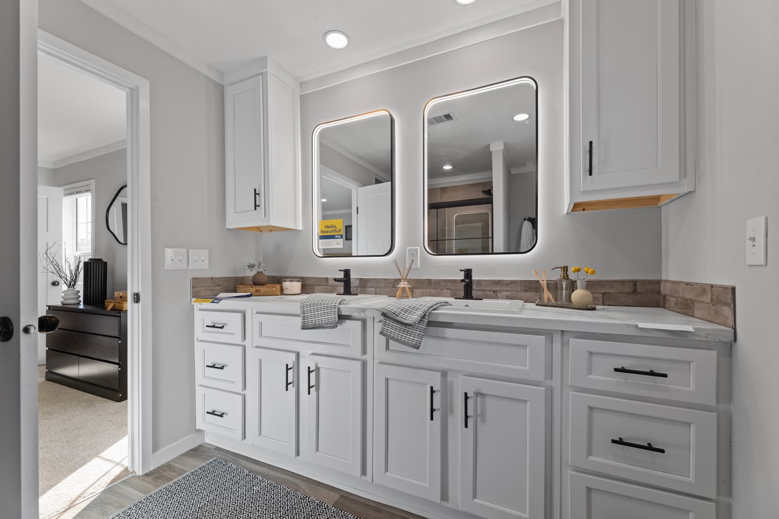 Bath 1. Double vanity with backlit mirrors, bedroom doorway at left.
