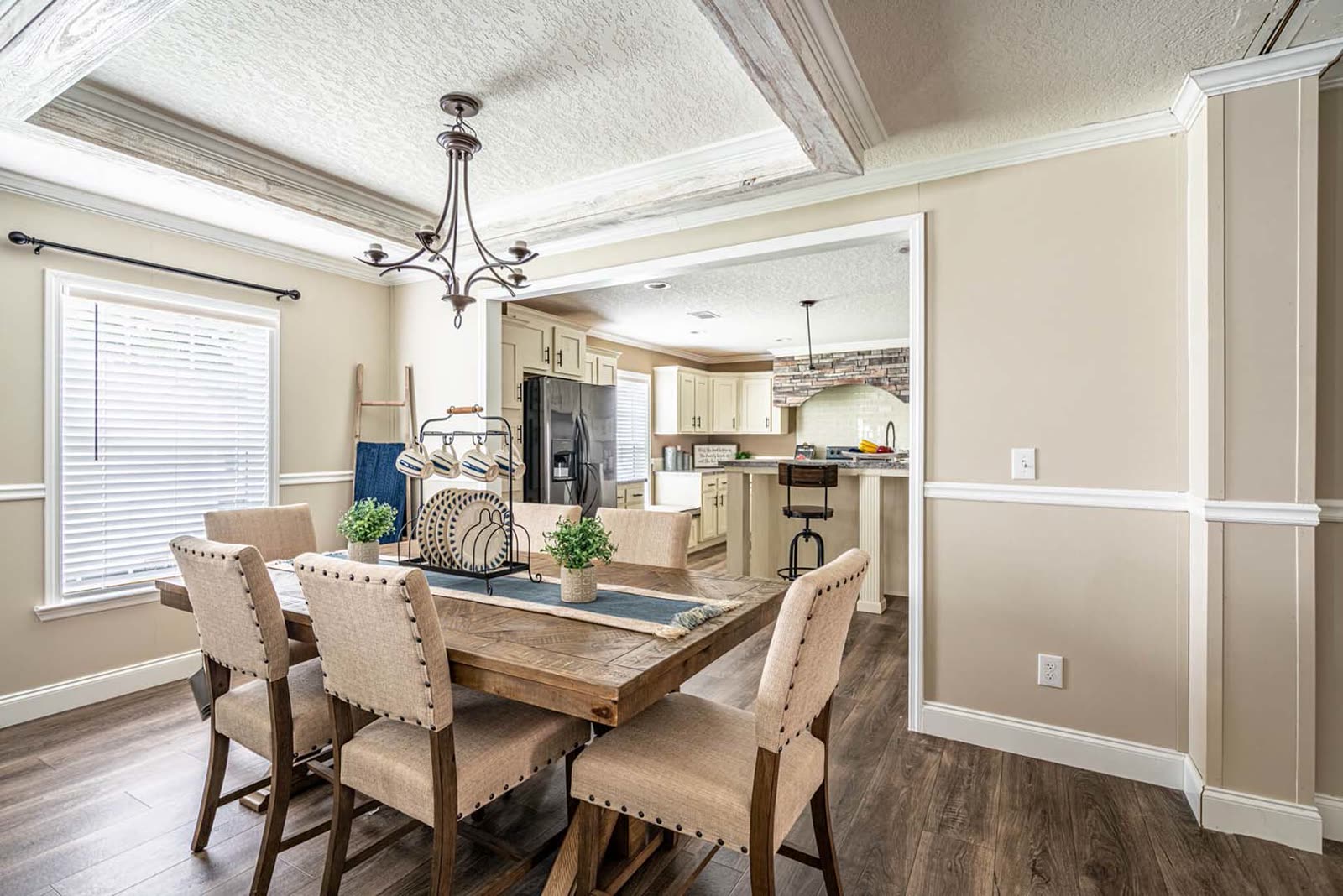 Dining room. Farmhouse table with upholstered chairs, chandelier, coffered ceiling, kitchen beyond.