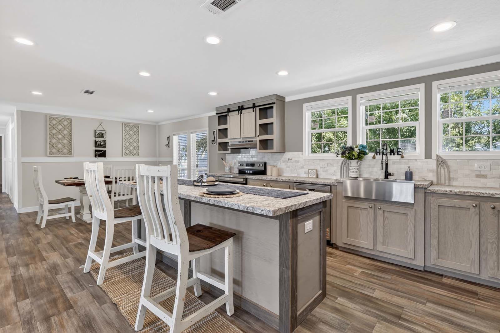 Kitchen. Grey cabinets, island with three stools, farmhouse sink, dining beyond.