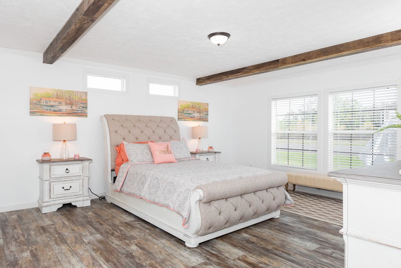 Primary bedroom. Upholstered bed, nightstands, windows on two walls, beam ceiling.
