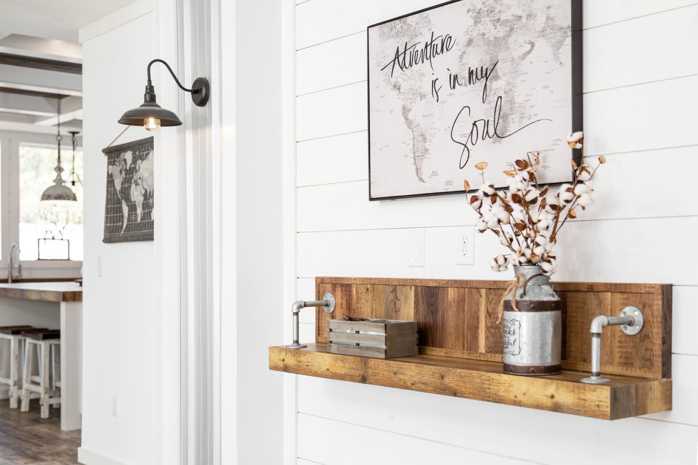 Living room. Wood shelf and gooseneck sconce on the shiplap wall, kitchen through the doorway.