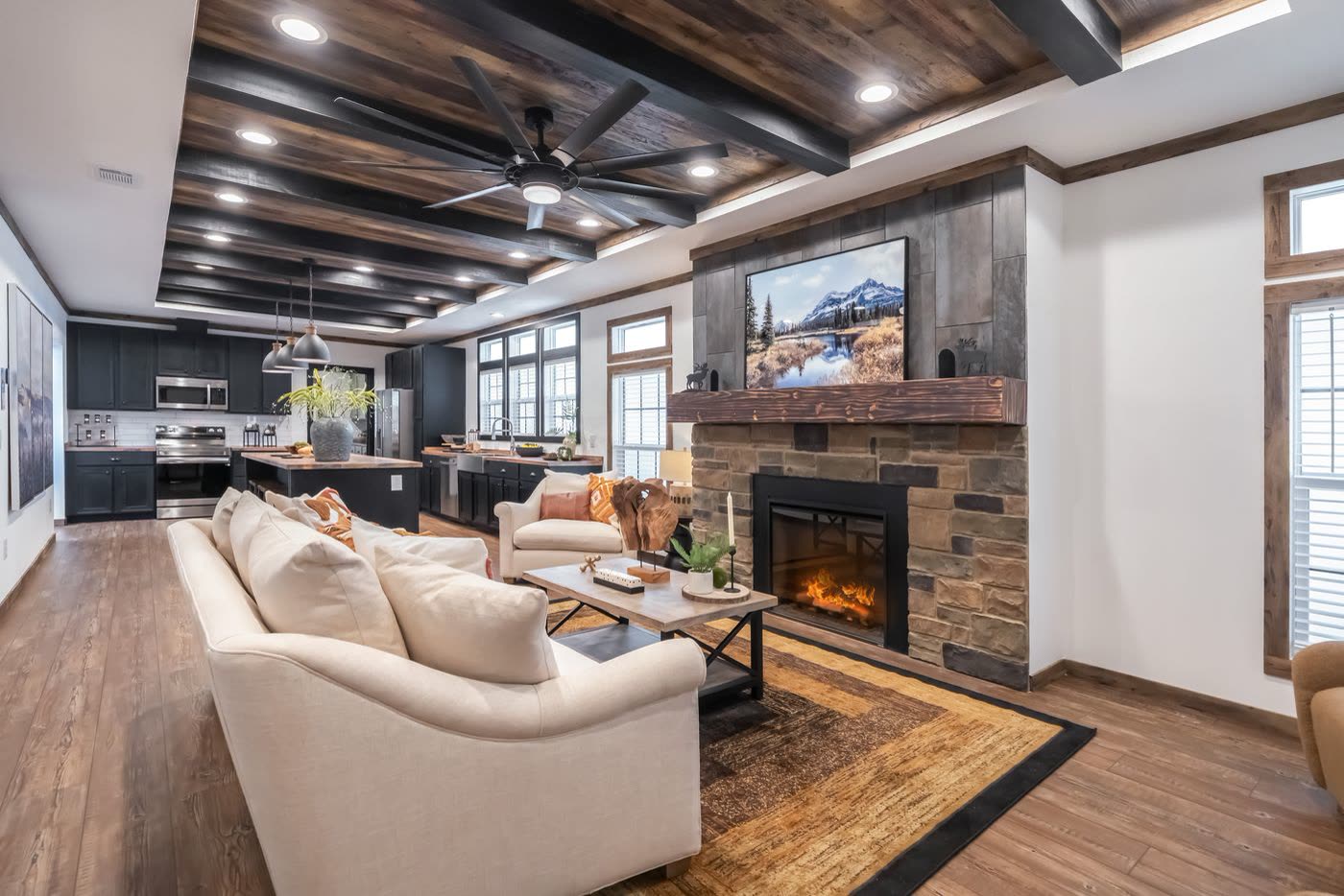 Living room. Two cream sofas, stone fireplace with television, beamed ceiling, kitchen beyond.