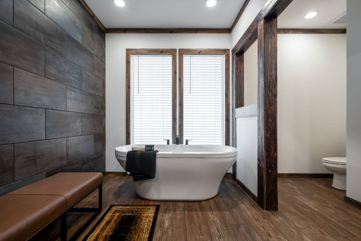 Primary bath. Freestanding tub under the window, leather bench, dark tile wall.