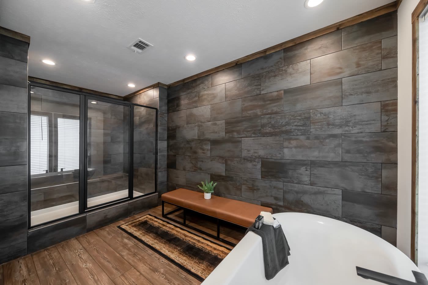 Primary bath. Tub room with dark tile wall, glass shower at left, bench.